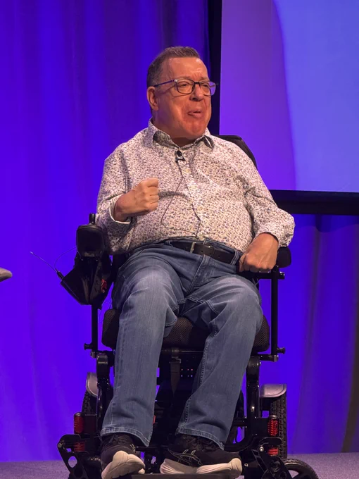 Here’s the alt text:

Dave Dame speaking on stage, seated in his power wheelchair, gesturing with a raised fist while mid-sentence. He is wearing a floral button-up shirt and jeans against a vibrant purple backdrop.