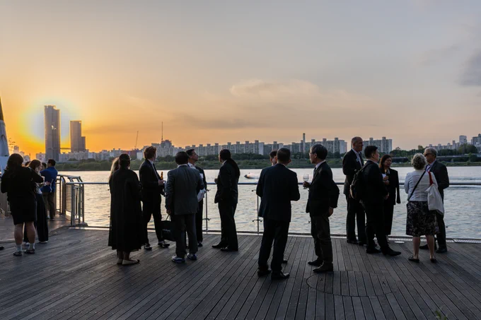 A group of people in formal attire stand on a wooden deck overlooking a river, engaging in conversation as the sun sets in the background.
