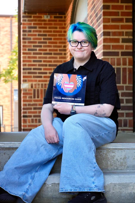 Recent MSLIS graduate Mads Christiansen smiles atop the staircase at the iSchool building while holding the 2026 edition of the Trans Resources Guide. 