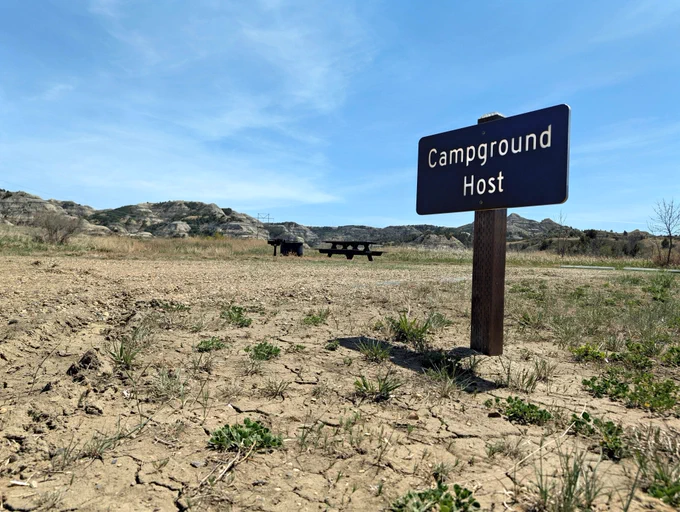 A Campground Host sign stands beside a wide, open campsite equipped with a picnic table, fire ring and grill. The site is reserved for the volunteer host who stays on location Thursday through Monday each week to assist visitors and answer questions. Forest Service photo by Scott C. Marvin