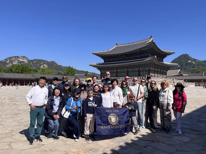 A group of people posing in front of Gyeongbokgung Palace in Seoul. The sky is clear, and they hold a banner reading 'Welcome Alumni Association.' The historic architecture and mountains are visible in the background.