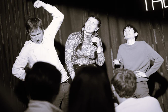 Three people onstage. One raises their arm above their head as they look down at something to adjust or smash. Two people stand behind them. Their focus is on the person's raised arm.
