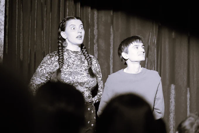 Two people stand onstage with straight backs and their arms behind their backs. One has a curled look and a snarled expression. The other has wide open eyes, a shocked expression and a slightly open mouth.