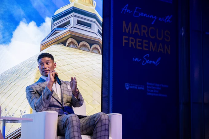 A person wearing a checkered suit sits on a white chair, speaking into a microphone. Behind them, a large screen displays an image of a golden dome with blue sky and white clouds.