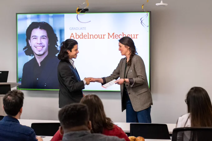 A graduate receives a certificate from a presenter in front of an audience during a ceremony with the graduate's name and photo displayed on a screen.