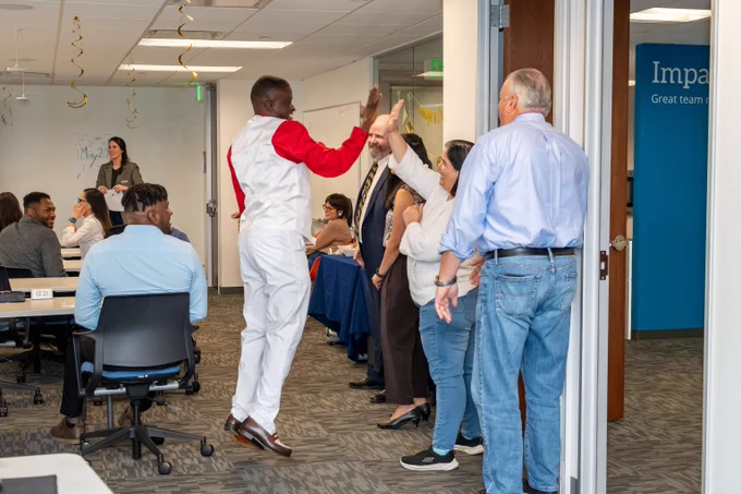 Man in white and red outfit jumps to high-five a colleague in an office filled with seated and standing coworkers.
