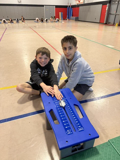 Two young boys sitting on the floor of an indoor gymnasium, looking at a blue exercise equipment.