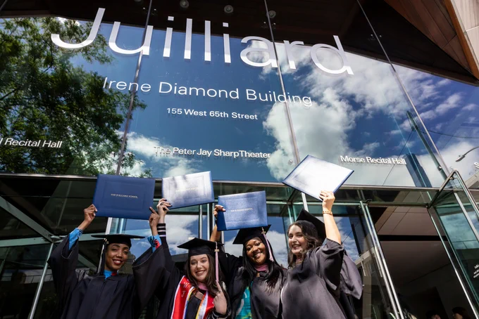 Graduates in caps and gowns hold diplomas above their heads outside the Juilliard School's Irene Diamond Building. A bright blue sky and clouds are reflecting in the glass of the building.