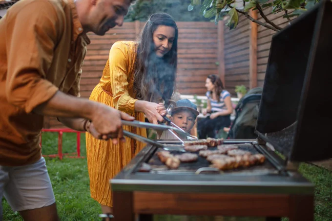 A man, woman, and child look on as food is cooked on a grill. 