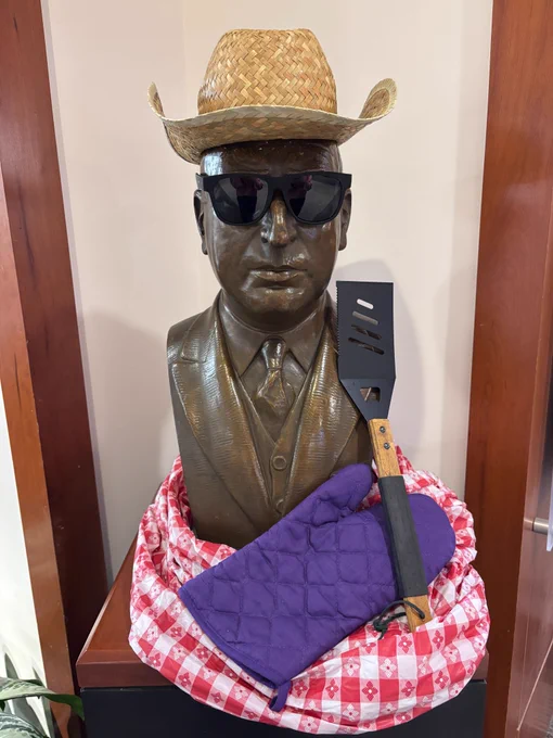 A bust of Arthur J. Morris wearing a hat and sunglasses. A tablecloth, oven mitt, and grilling spatula are on the pedestal.