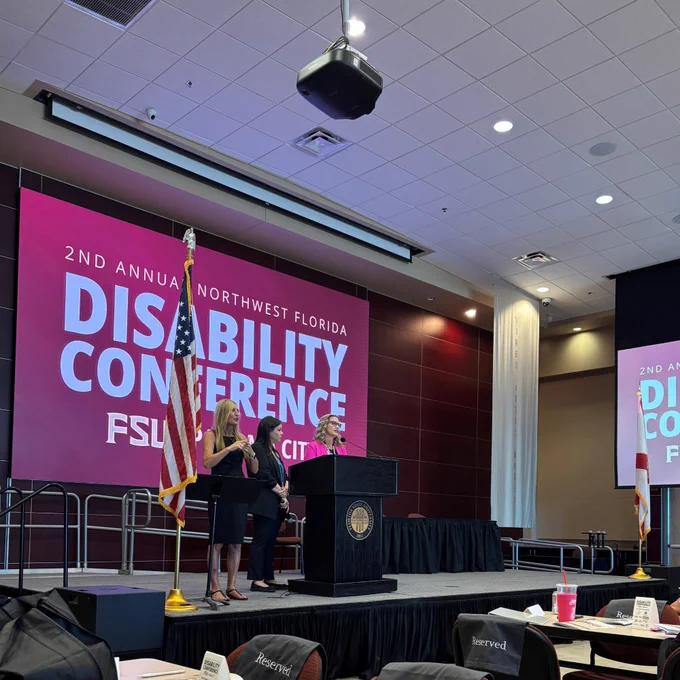 A large room with multiple projection screens that read "2nd Annual Northwest Florida Disability Conference, FSU Panama City." There are three individuals on a stage near a podium looking out over an audience seated at tables.