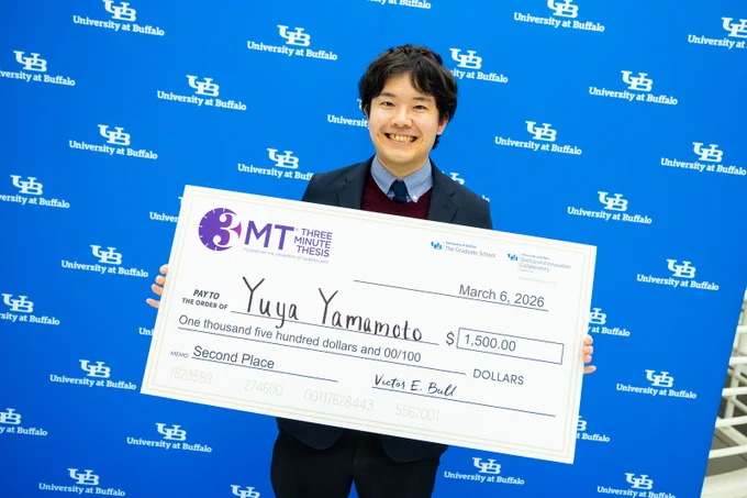 Yuya smiling and holding an oversized check for $1,500.00 made out to \"Yuya Yamamoto\" for \"Second Place\" from the \"3MT Three Minute Thesis\", dated March 6, 2026. The background has logos of \"University at Buffalo\".