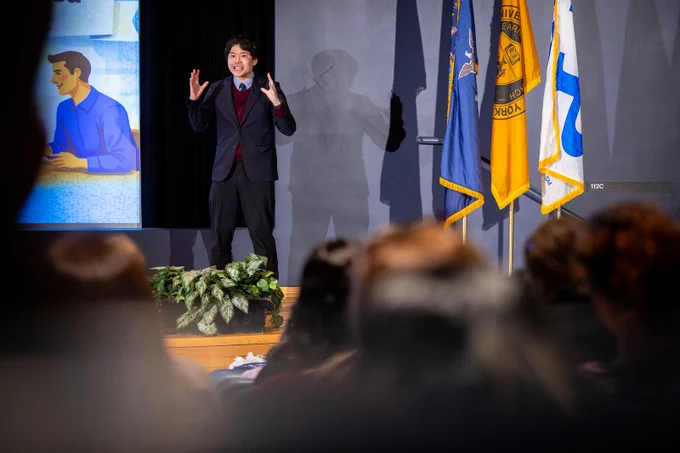 On stage, a speaker with dark hair gestures, wearing a suit and sweater. Behind, a screen shows a cartoon man and flags stand nearby. An audience watches from below the stage. \"112C\" is visible on the wall.