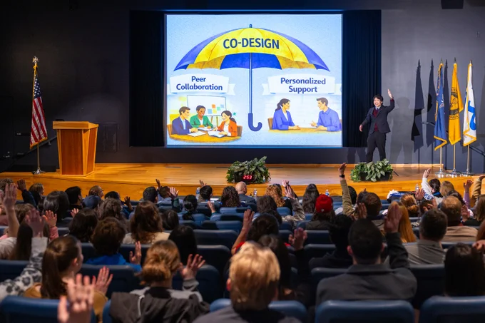 Audience with raised hands views a presentation on \"CO-DESIGN\", symbolized by an umbrella covering \"Peer Collaboration\" with people at a table, and \"Personalized Support\" showing two people talking. Presenter stands on stage with flags on his right.