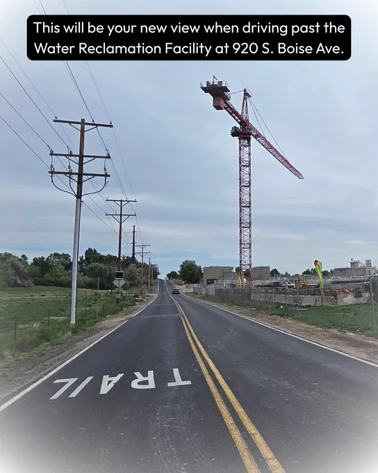 A two-lane road with "TRAIL" painted on it leads to a construction area at the Water Reclamation Facility on 920 S. Boise Ave. A large red crane and utility poles are visible along the road. A tree-lined area is on the left, and the sky is cloudy. Text reads: This will be your view when driving past the Water Reclamation Facility at 920 S. Boise Ave. 