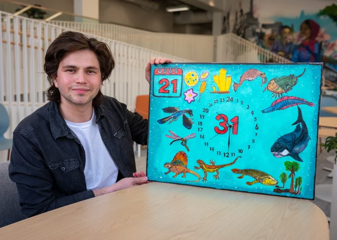 UTSC student holding a colorful calendar featuring various wildlife, including a whale and dinosaurs, with numbers in a circle. A colorful mural is visible in the background.