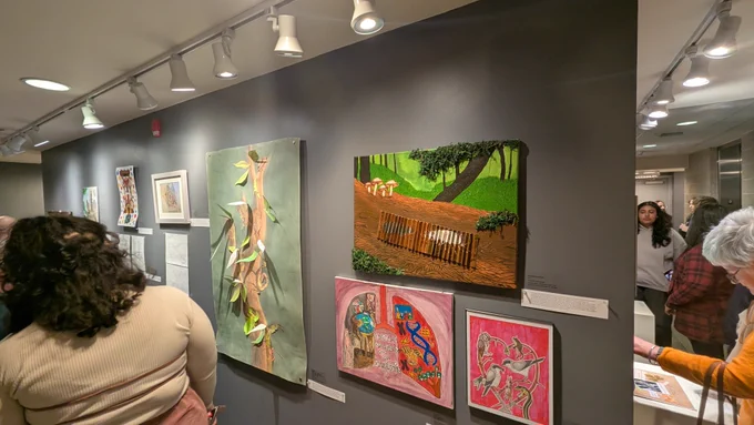A group of people viewing an Deep Time – Creative Minds exhibit in Station Gallery. Various artworks are displayed on a gray wall, including a painting of a forest scene with a wooden fence and colorful abstract pieces. The lighting is focused on the art, and attendees are engaged, observing the pieces.