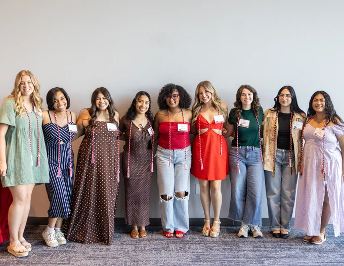 Nine women stand in a row against the wall with arms linked while wearing their honor cords to pose for a photo at the TSA graduation celebration