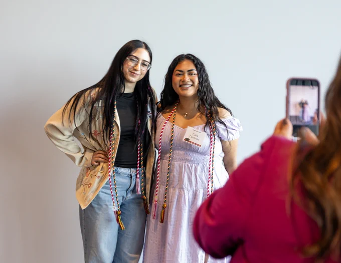 two students pose together for a photo wearing their honor cords while someone takes a photo of them on a cell phone