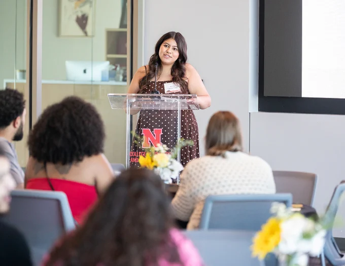 Jenny Castor Cruz speaks from behind a clear lectern as others listen to her remarks at the TSA graduation celebration
