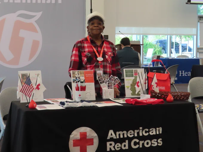 A Red Cross volunteer stands at a booth covered in a black tablecloth with the Red Cross logo. There are various flyers and Red Cross branded items on the table.