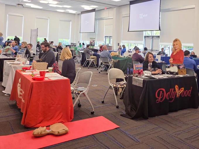 A wide shot of an indoor community fair. In the foreground there is a CPR mannequin on the floor and booths with red and black tablecloths.