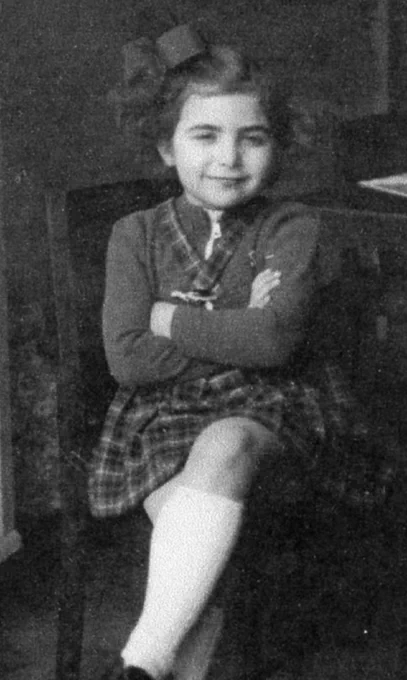 Black and white photo of a young girI seated with crossed legs, smiling, wearing a plaid dress and a bow in her hair.
