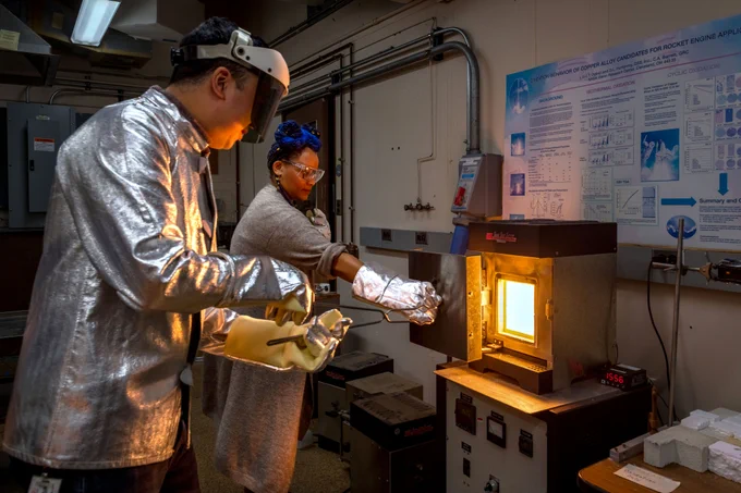 Two researchers wearing shiny, silver-colored protective gear including thick gloves, coats, and eyewear stand beside a furnace inside a laboratory as they prepare to remove a sample of a new material. The researcher on the left holds large tongs and reaches toward the furnace as the researcher on the right holds open the furnace door. The inside of the furnace is glowing bright white-orange. Credit: NASA/Jef Janis