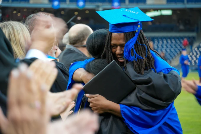 A person in a blue graduation cap and gown is joyfully hugging another person in similar attire at a graduation ceremony. People are clapping around them, and a stadium with rows of seats is visible in the background.