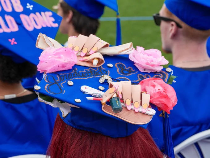 A decorated graduation cap in a crowd features the word "Graduated" and is adorned with artificial nails, pink flowers, and makeup items.