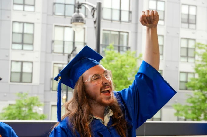 A person wearing a blue graduation cap and gown is raising a fist in celebration. They are standing outside with a building and trees in the background, expressing excitement and joy.