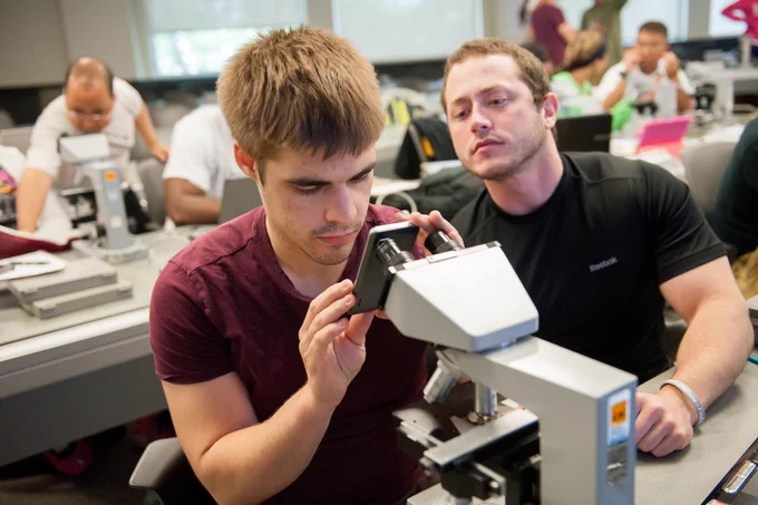 A young man looks intently through the lens of a microscope in a laboratory classroom. Another man sits next to him, closely observing the workspace.