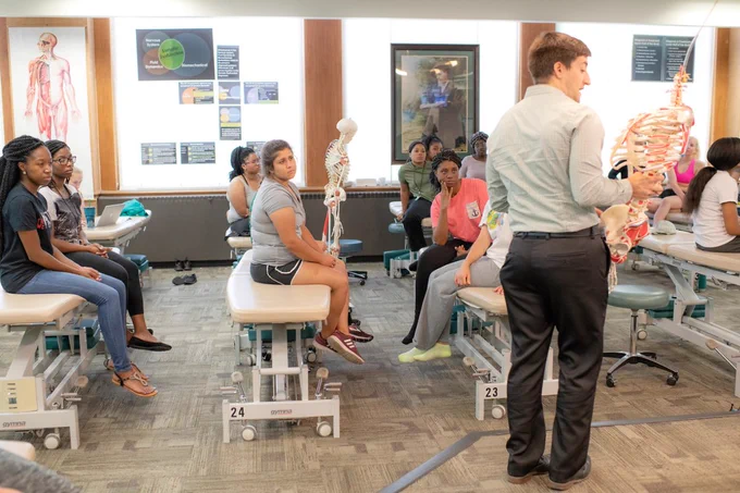 An instructor stands in an osteopathic manipulative medicine lab holding an anatomical model of a human spine and pelvis. A group of students sit on OMM tables, listening attentively to the demonstration.