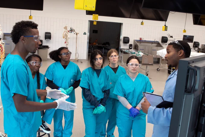 A group of students wearing teal scrubs and safety glasses gather in a laboratory setting. They listen closely to an instructor wearing a light blue lab coat who is gesturing and holding a tablet.