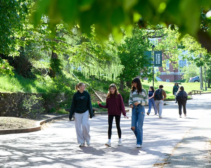 A group of people walking on a sunlit path surrounded by trees. A banner in the background reads "Welcome Home.