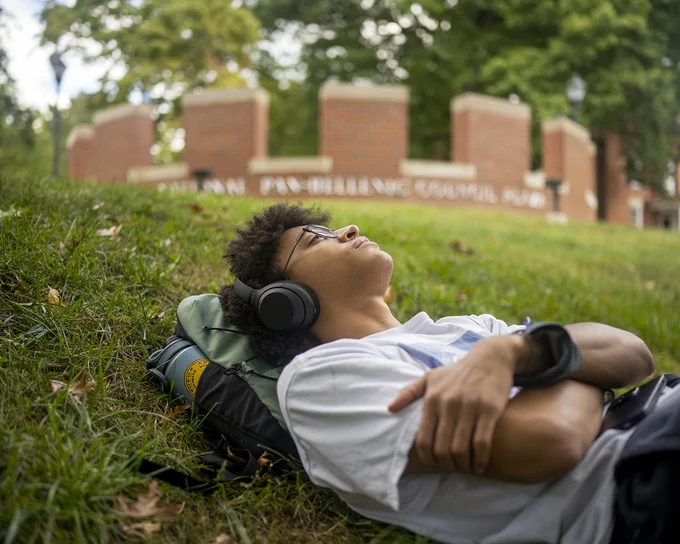 A person is lying on the grass with headphones, eyes closed, and head resting on a backpack. Behind them, a brick sign reads "National Pan-Hellenic Council" amidst lush greenery.