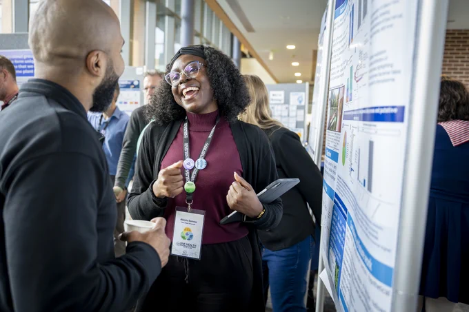 A person is engaging in a lively conversation at a conference or event. They are holding a tablet and wearing a lanyard with identification. Around them, others are interacting and viewing posters with charts and text.