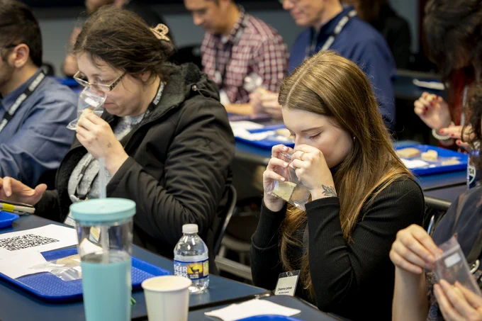 A group of people seated at tables are engaged in a sensory evaluation session, smelling samples in clear plastic bags. Each table has a blue tray, a small water bottle, and plastic bags containing bread samples. Participants appear focused on the activity.