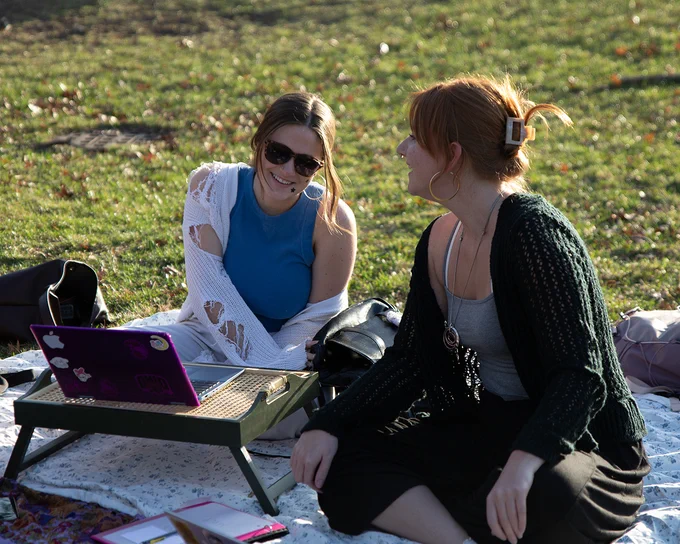 Two people are sitting on a blanket in a grassy area, enjoying a sunny day. A laptop with stickers on it is on a small portable table. Both individuals appear relaxed and engaged in conversation.