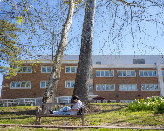 A person sits on a wooden bench reading a book beneath large trees. In the background, a brick multi-story school building is visible against a clear blue sky.