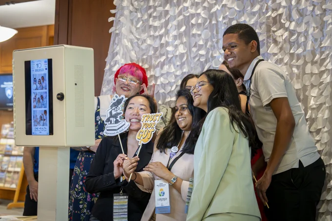 A group of people are gathered around a photo kiosk, enjoying themselves. One person holds a sign that says "Trust Your Gut." There's a textured, decorative backdrop behind them. They appear to be at a social event or gathering, with cheerful expressions and a relaxed atmosphere.