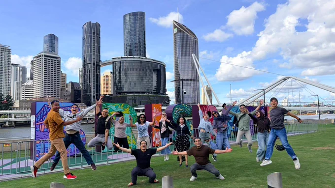 Group photo of participants jumping and posing joyfully in front of the 'Brisbane' sign, in a city park with modern skyscrapers and a bridge in the background under a blue sky.