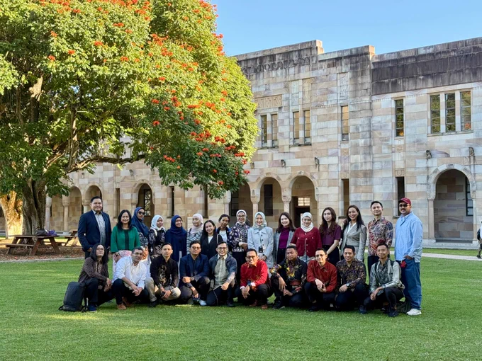 Group photo of participants posing on the green lawn of The University of Queensland Great Court in front of historic stone building under a large tree with orange flowers.