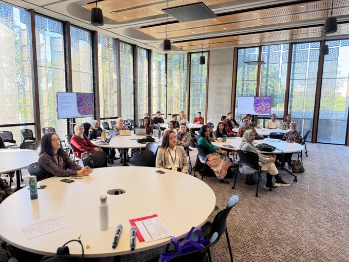 Participants listening to a presentation in a modern classroom setting with large windows and presentation screens during a short course session.