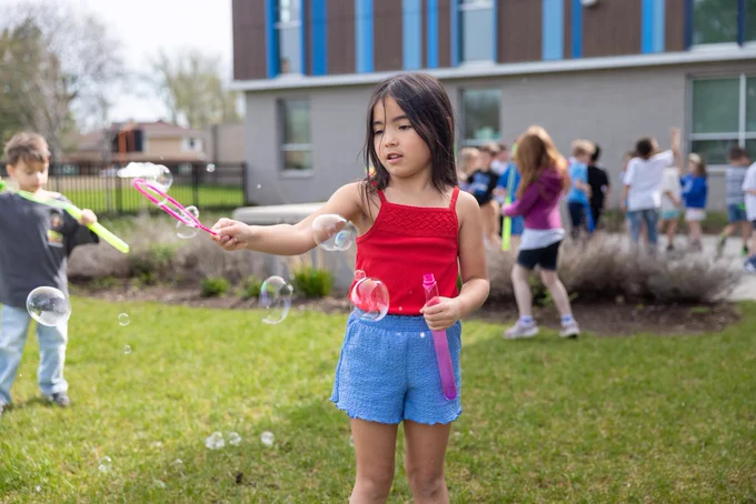 A group of children enjoy a sunny day outside, playing with bubbles. The kids are engaged and having fun on a grassy area near a building.