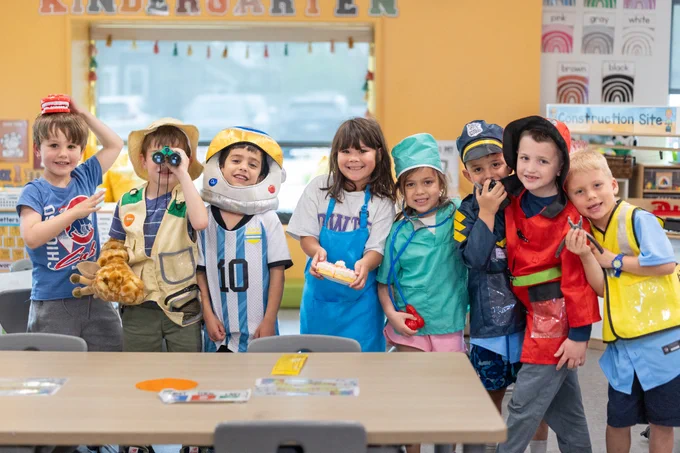 A group of children in colorful dress-up costumes pose happily in a kindergarten classroom. The setting is lively and playful.