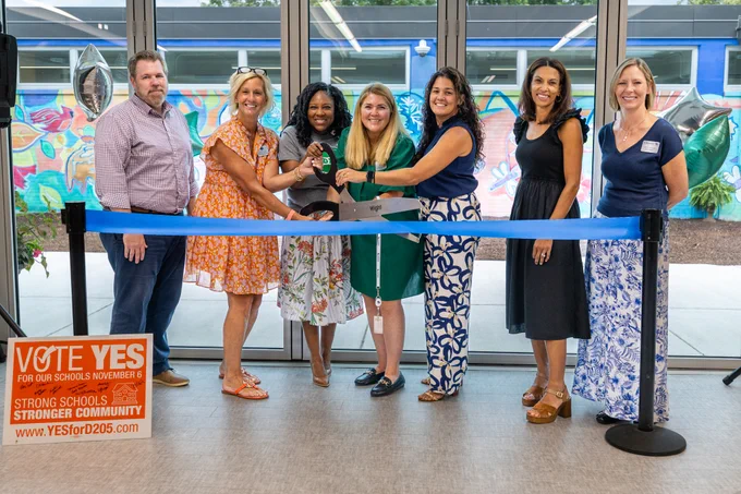 A group of people at a school ribbon cutting ceremony with a 'Vote Yes' sign. They smile and prepare to cut the ribbon together.