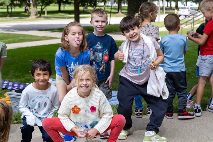 A group of cheerful children pose playfully outdoors. The sunny day highlights the lively joy and energy of the event.
