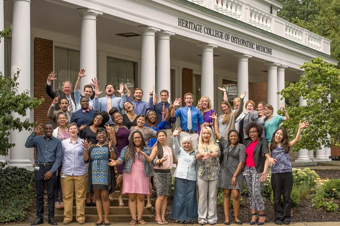 A group of young adults and faculty wave and pose excitedly on the steps of a brick building with white columns. Above them, a sign on the building reads "Heritage College of Osteopathic Medicine."