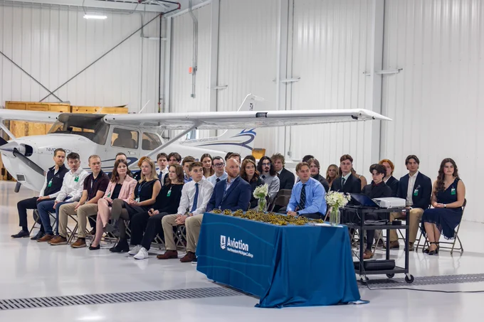 A group of people is seated in a spacious hangar with an airplane parked in the background. In front of them, there is a table covered with a blue cloth featuring a logo that includes the word "Aviation." The setting suggests a formal event or ceremony.